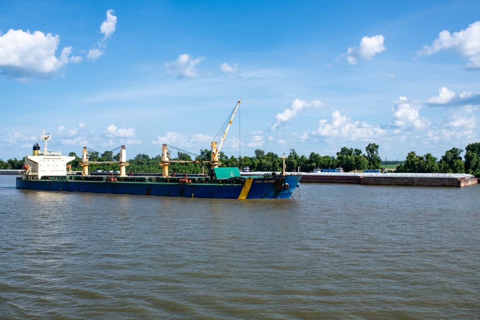 Exportação Brasileira. — A cargo ship navigates the Mekong River surrounded by a lush green forest under a clear blue sky.